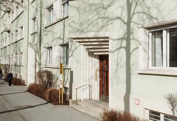 Eingang eines Mehrfamilienhauses mit grüner Fassade und Schatten von Bäumen im Sonnenlicht.